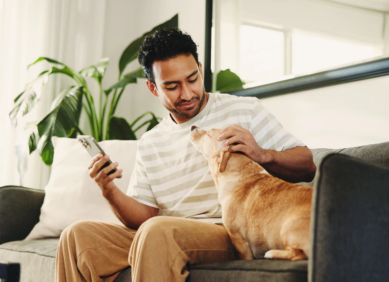 A man sitting on a couch, smiling while holding a smartphone and petting a light brown dog. The room has bright natural light and green plants in the background.